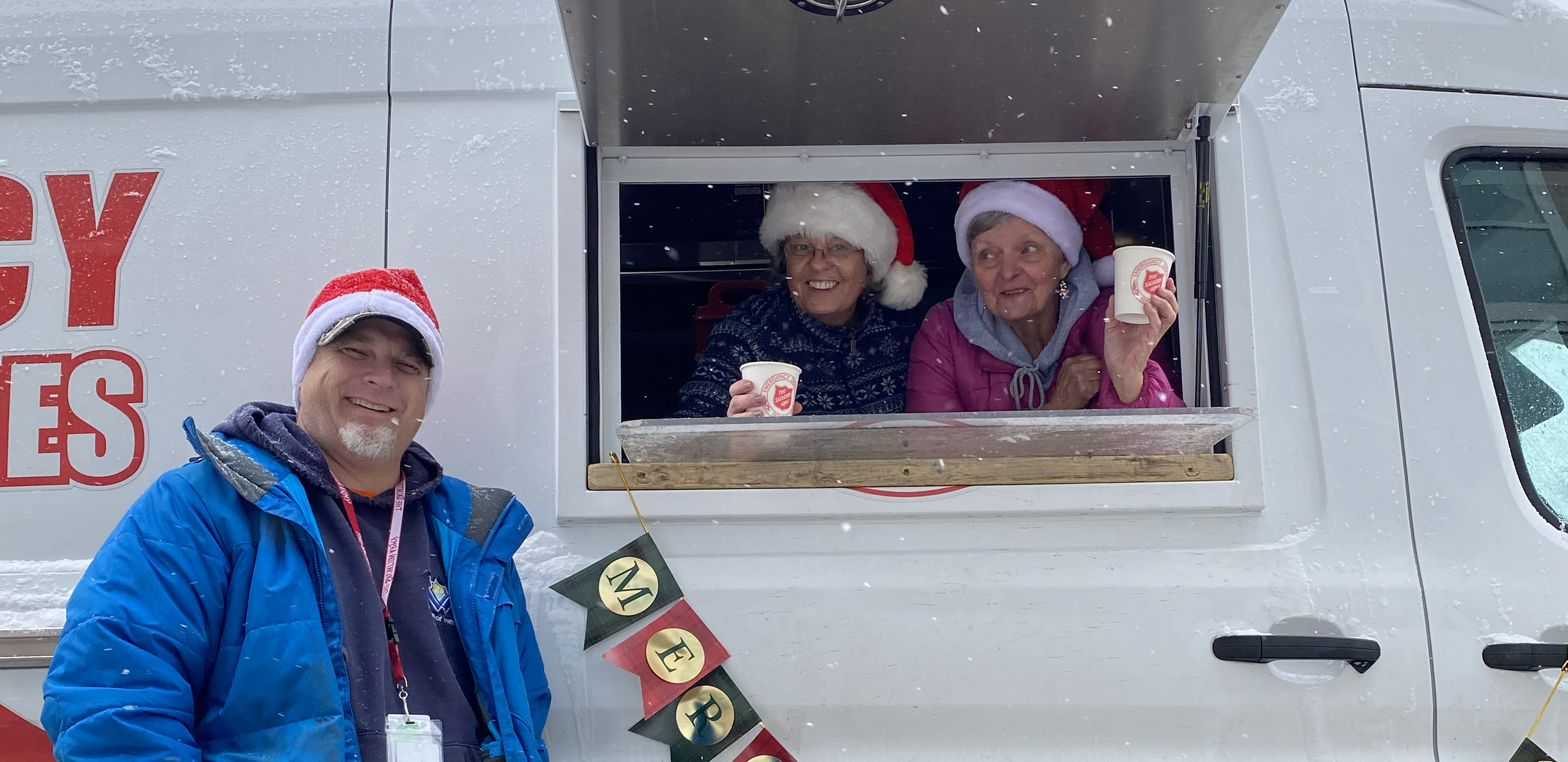The Salvation Army Emergency Disaster Services truck with a serving window open, where people inside the vehicle hand out hot drinks while a volunteer stands outside, with light snowfall visible and holiday decorations including Santa hats and a small festive banner.